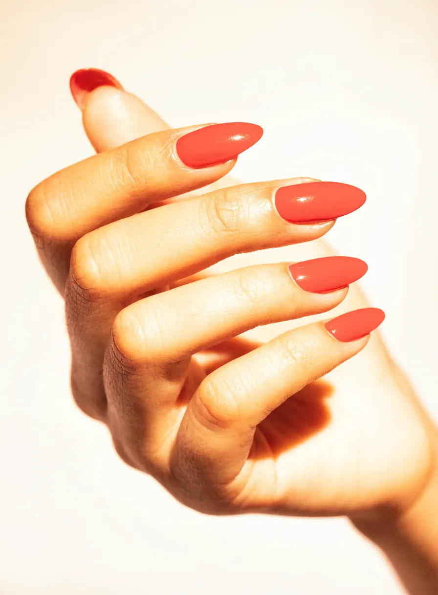 Close-up of a hand with glossy coral-red almond-shaped nails against a cream background
