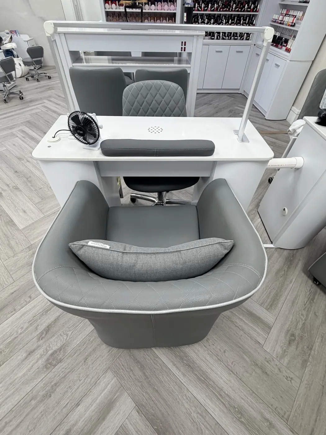 A manicure desk with a white top, gray client chair and lamp, with polish shelves behind it