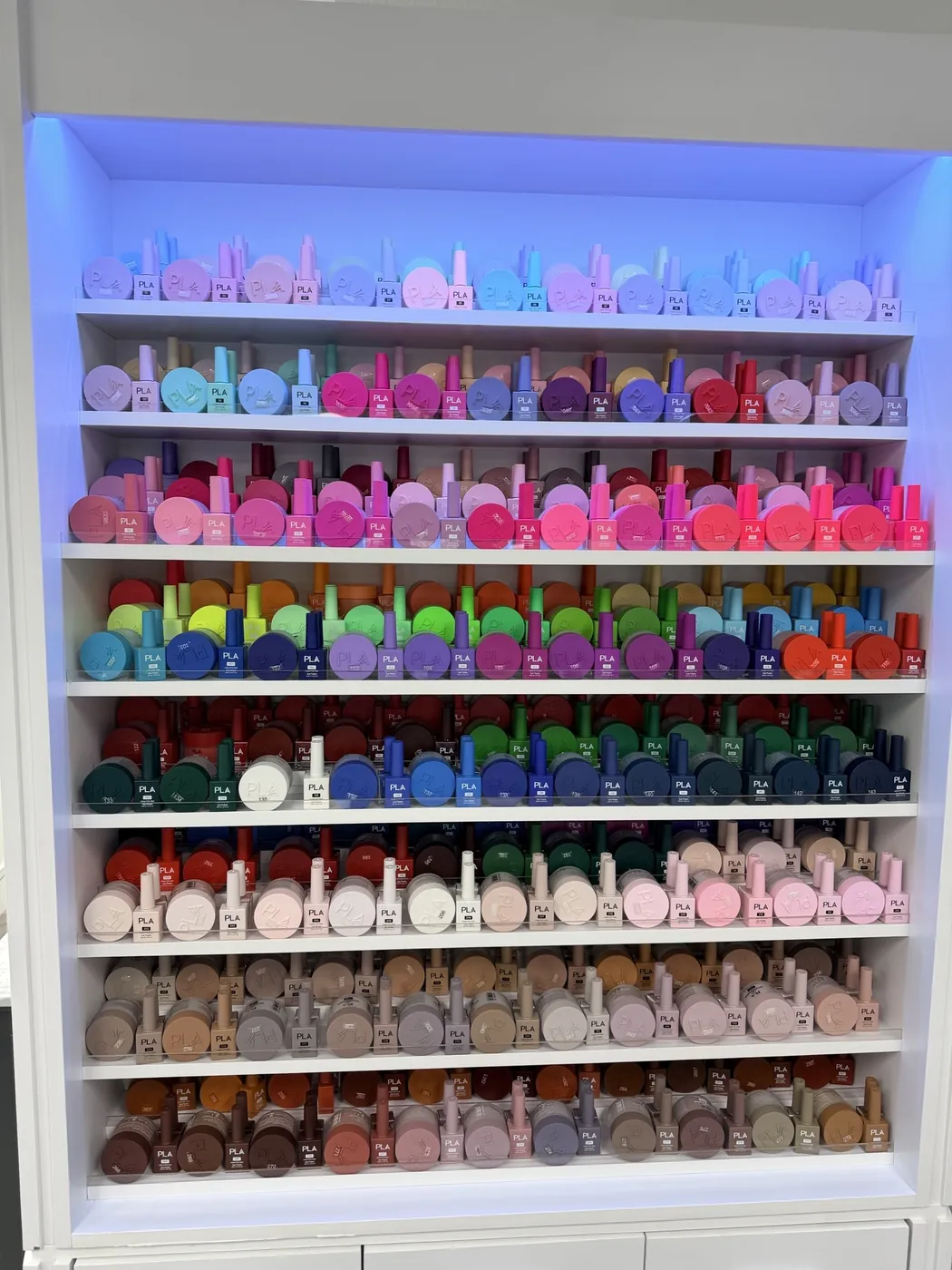 Floor-to-ceiling shelves of nail polish bottles arranged by color and lit from behind at Legend Nail Studio